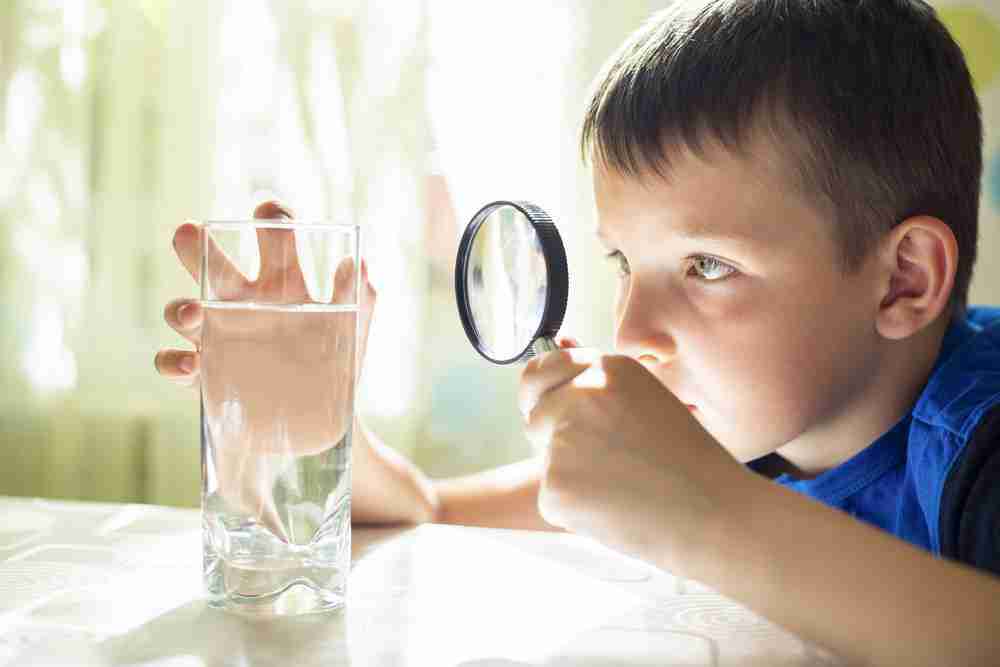 A child inspecting a clear glass of water using a magnifying glass.