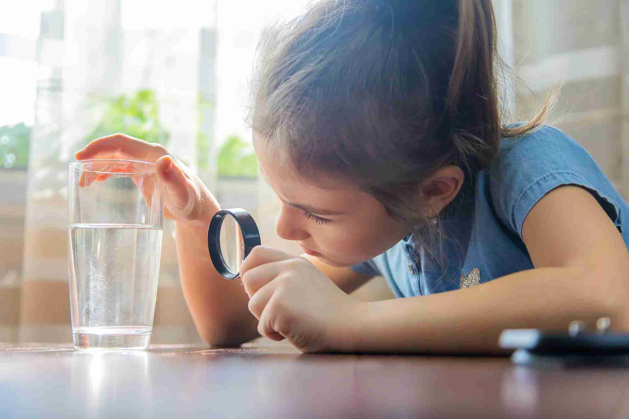 A young girl using a magnifying glass to inspect a clear glass of water.